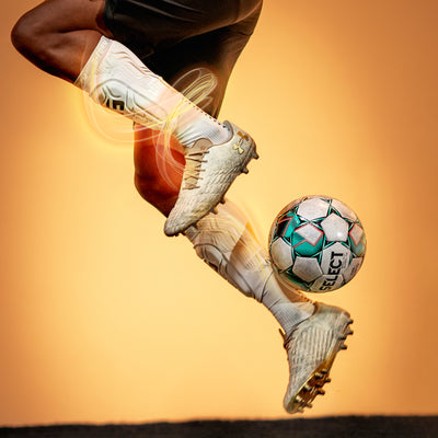 Soccer player's feet in white cleats kicking a soccer ball with a yellow background