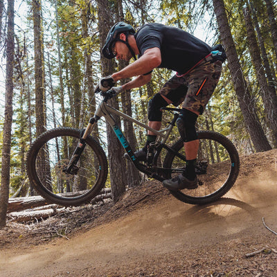 Mountain biker riding through a forest trail, wearing a USWE Zulo Hydration Waist Pack. The rider is mid-action, lifting the front wheel of their bike on a dirt path while sporting protective gear, including a helmet and knee pads. | 8Lines Shop